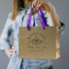 A medium brown paper bag with purple ribbon handles, featuring a logo on the front.