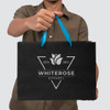 A person holds a large black paper bag with a blue ribbon handle and a logo on the front.