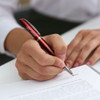 A red metal pen is being used to write on a sheet of paper, with a person's hand visible.