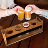 A wooden taster tray with five circular cutouts holding glasses of beer and small bowls. The tray has a rustic finish.