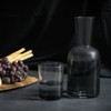 A glass carafe and tumbler set in smoky grey, accompanied by grapes and breadsticks on a wooden platter.