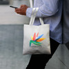A cream tote bag with a colourful logo, featuring sturdy handles, being held by a person outdoors.