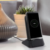 A wireless charging station in black with a smartphone displaying a charging indicator, set near a decorative plant.