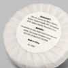 A white circular soap wrapped in paper, labelled with ingredients and warnings, features a logo.