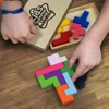 A colourful wooden pentomino puzzle set displayed on a wooden surface, with hands assembling the pieces.