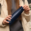 A drink bottle in navy blue, held by a person wearing a beige jacket, featuring a logo on the side.