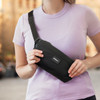 A black sling bag is held by a person wearing a light purple T-shirt, with a logo visible on the bag.