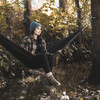 A woman in a beanie and flannel sits in a black outdoor hammock among autumn trees.