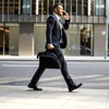 A man in a suit carries a black laptop conference bag while walking down a city street. The bag has a logo.