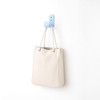 A cream-coloured tote bag hanging on a blue hook against a white wall. The bag has a simple design and a logo.