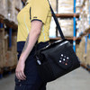 A black lunch cooler bag with a shoulder strap, featuring a logo, shown on a person in a warehouse setting.