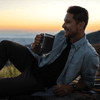 A thermal copper mug in matte grey held by a person sitting outdoors against a picturesque sunset backdrop.
