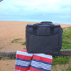 A grey cooler bag made from recycled material, placed on a wooden railing beside a beach towel.