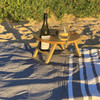 A wooden picnic table in natural tones on sand, holding a wine bottle and glass, with a checkered and striped blanket.
