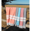 A collection of four Turkish towels in coral, blue, grey, and beige draped over a wooden railing against a beach backdrop.