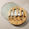 A round bamboo cheese board set featuring four stainless steel knives and a corkscrew, with a glass lid.