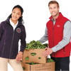 A man in a red vest stands beside a woman in a dark purple vest, both holding boxes filled with leafy greens.