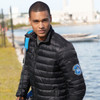 A man wearing a black puffer jacket stands by a waterway, with a logo on the sleeve.