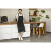 A black apron is worn by a man in a modern kitchen, featuring wooden accents and plants. The setting is bright and contemporary.