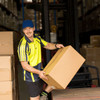 A man in a high-visibility yellow and black shirt and blue cap lifts a cardboard box in a warehouse setting.