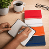 A set of colourful notebooks in red, white, orange, and blue, with a hand writing in a black notebook. A coffee cup and glasses are nearby.