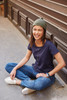 A smiling woman in a navy T-shirt and green beanie sits casually on a path, wearing jeans and white shoes.