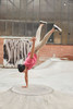 A person performing a handstand in a pink T-shirt and beige pants at a skate park with industrial background.