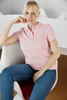 A light pink polo shirt displayed on a model seated in a modern chair, featuring a collar and a logo.