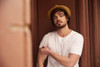 A man wearing a plain white T-shirt and a straw hat, posing against a brown background.