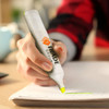 A highlighter being held by a person, featuring a green tip and a branding logo, with writing on paper in the foreground.