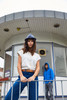 A woman in a white casual T-shirt and blue pants poses in front of a building, wearing a grey hat. A person in a blue hoodie is in the background.