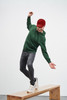 A man wearing a green hooded sweatshirt and a red cap balances on a wooden bench, dressed in grey jeans and white sneakers.