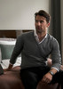 A man wearing a grey knitted jumper sits on a bed, with a watch on his wrist and a neutral-toned room in the background.