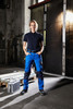 Workwear pants in blue and black, worn by a person standing in an industrial setting. The outfit includes a black polo shirt.
