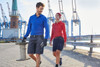 A blue long-sleeve polo shirt and a red polo shirt are worn by two models in a dockyard setting. Both have logos.