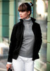 A women's black jacket with a high collar, worn over a grey sweater and white pants, featuring a logo.