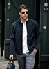 A man in a black soft shell jacket walks confidently, wearing sunglasses and holding a bag.