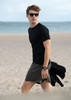 A young man wearing a black T-shirt and grey shorts walks on a beach, with sunglasses and a watch.