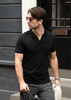 A man in a black V-neck polo shirt and sunglasses walking outside, carrying a bag and a hat.