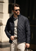 A navy quilted men's jacket features a zip front and a fitted design, worn with a light sweater and beige trousers.