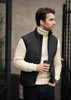 A black men's vest is worn over a cream cable-knit sweater, paired with dark jeans and holding a coffee cup.