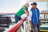 A woman in a green vest and a man in a blue vest smile on a boat deck with scenic views in the background.