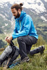 A blue men's outdoor jacket is worn by a person kneeling on grass next to a backpack in a mountainous landscape.