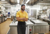 A man in a yellow polo shirt and black apron stands in a clean, modern kitchen, smiling confidently.
