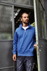 A blue half zip sweat jumper with a collar, worn by a man standing at a doorway, featuring a logo on the chest.