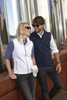 A white softshell vest and a navy softshell vest are worn by two people standing next to metal structures.