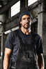 A men's workwear T-shirt in black, featuring a plain design, worn under black overalls in an industrial setting.