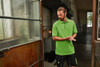 A green men's polo shirt is displayed, worn by a man standing near an industrial door. The shirt features a logo.