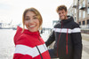 A red and black soft shell jacket with white stripes, worn by a smiling woman and a man by the water.
