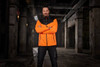 An orange and black men's allweather jacket is displayed on a model in an industrial setting. The jacket has a logo.
